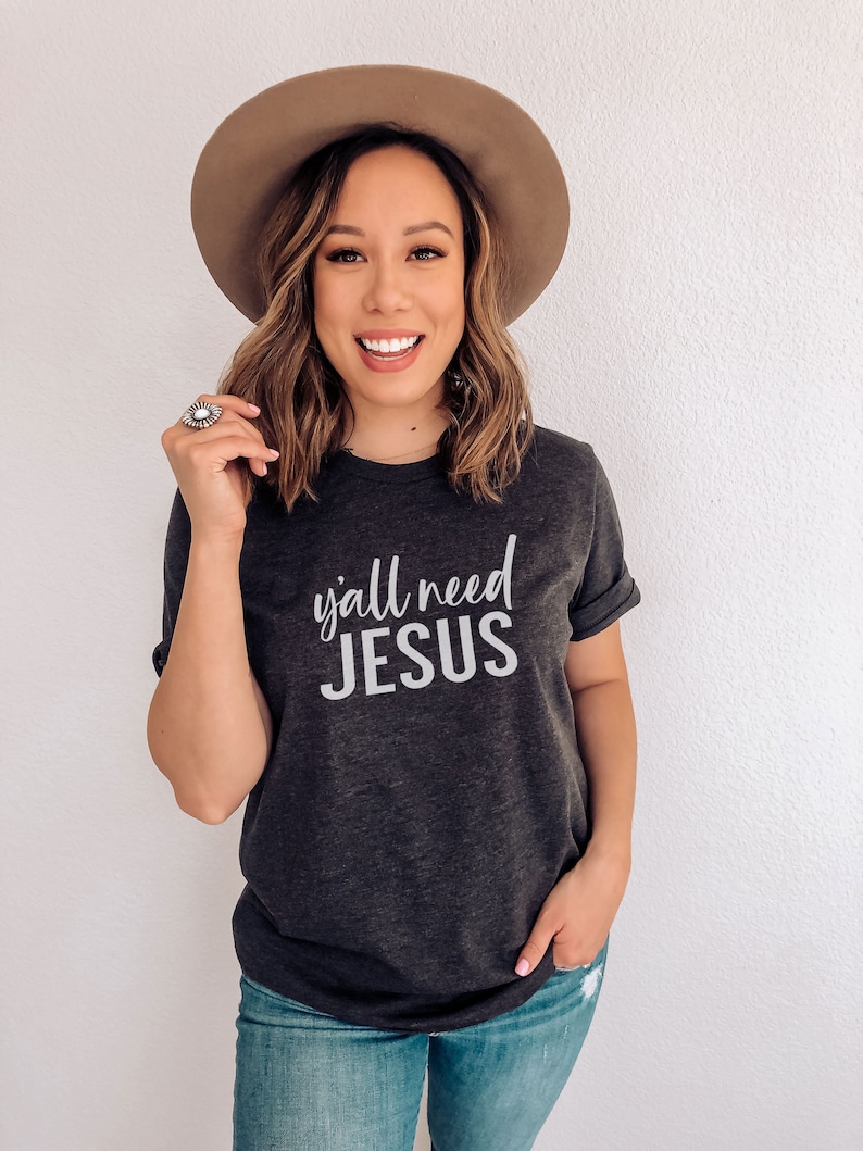 May include: A woman wearing a dark gray t-shirt with the text "y'all need JESUS" in white lettering. She is also wearing a brown hat and blue jeans.