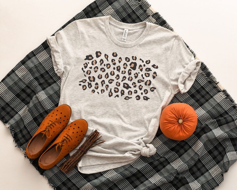 May include: A gray t-shirt with a brown and black leopard print design. The shirt is laying on a black and white plaid blanket with brown leather shoes and a small orange pumpkin.