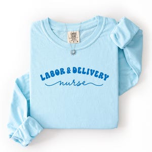 May include: A light blue long-sleeve shirt with the words "LABOR & DELIVERY nurse" in blue script. The shirt is folded, with the sleeves rolled up. A silver necklace with a sunburst pendant is on the shirt.