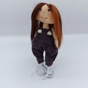 Puede incluir: Muñeca hecha a mano con cara de tela, cabello castaño y rojizo, y una sonrisa cosida. La muñeca viste un peto a cuadros morados con zapatillas blancas. La muñeca mide aproximadamente 15 cm de altura.