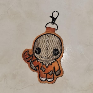 May include: A brown and orange plush keychain with a stitched face and a candy corn in its hand. The keychain has a silver metal clasp.