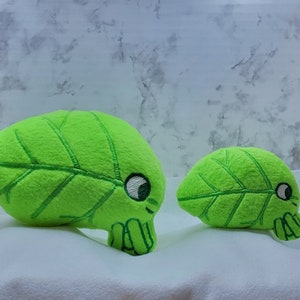 May include: Two plush green leaf-shaped toys with embroidered details and eyes. The larger toy is lying on its side, while the smaller toy is sitting upright.