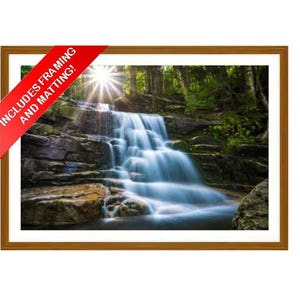 May include: A framed print of a waterfall cascading over rocks, surrounded by lush green trees. The image is in a brown wooden frame with a white mat. A red banner in the corner reads "INCLUDES FRAMING AND MATTING!"