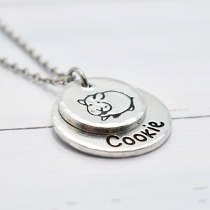 May include: Silver necklace with two round pendants. The top pendant features a small pig illustration. The bottom pendant has the word "Cookie" engraved on it.