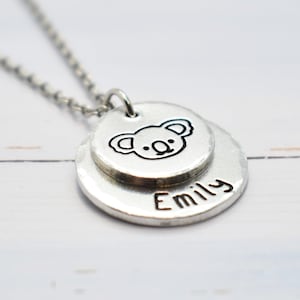 Könnte beinhalten: Eine silberfarbene Halskette mit zwei runden Anhängern. Der obere Anhänger zeigt eine Koala-Bär-Illustration, und der untere Anhänger trägt den Namen "Emily". Die Halskette hängt an einer filigranen Kette.