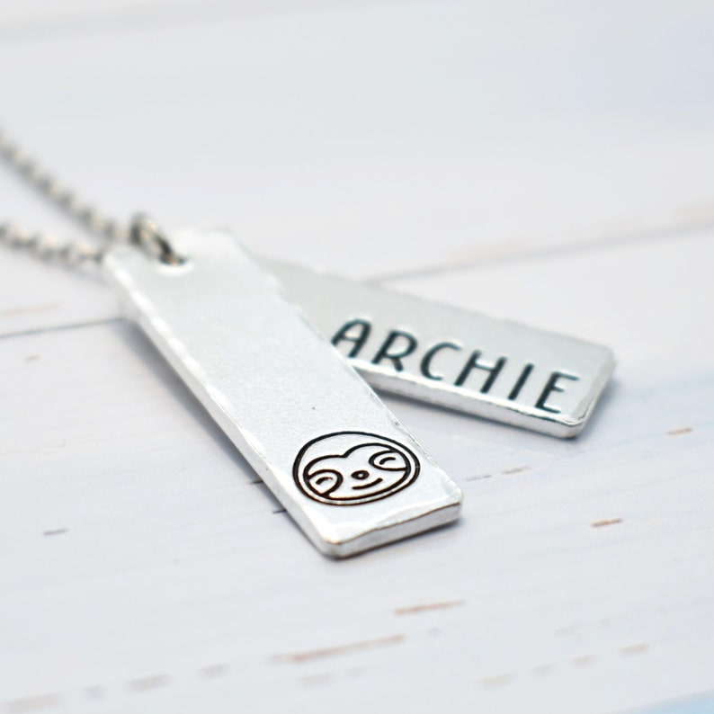 May include: Silver necklace with two rectangular tags. One tag has the name "ARCHIE" engraved on it. The other tag has a small sloth face engraved on it.