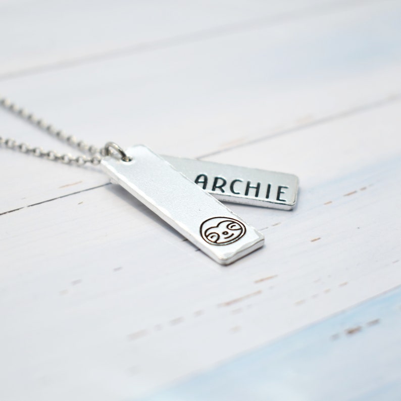 May include: Silver necklace with two rectangular tags. One tag has the name "ARCHIE" engraved on it. The other tag has a small sloth face engraved on it.