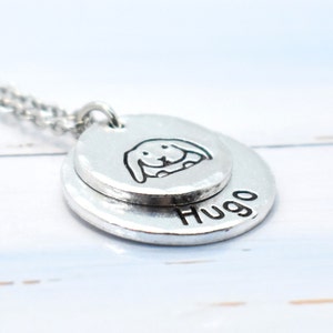 May include: Silver necklace with two round pendants. The top pendant has a small, hand-drawn image of a rabbit. The bottom pendant has the name "Hugo" engraved on it.