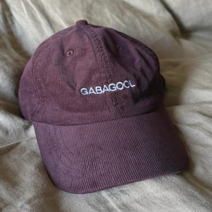 Puede incluir: Gorra de béisbol de pana burdeos con la palabra "GABAGOOL" bordada en blanco. La gorra tiene una visera curva y una corona estructurada. El sombrero descansa sobre una tela arrugada de color marrón claro.