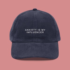 May include: Navy blue corduroy baseball cap with the white text "ANXIETY IS MY INFLUENCER" embroidered on the front. The cap has a curved brim and a rounded top.