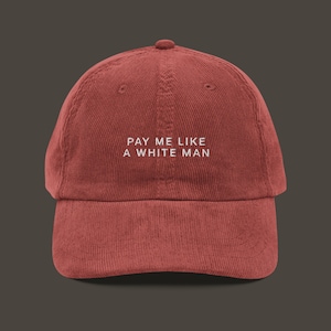 May include: A red corduroy baseball cap with the text "PAY ME LIKE A WHITE MAN" embroidered in white.