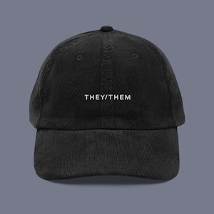 May include: A black corduroy baseball cap with the text "THEY/THEM" embroidered in white on the front.