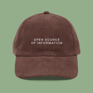 May include: A brown corduroy baseball cap with the text "OPEN SOURCE OF INFORMATION" in white. The cap has a curved brim and a rounded top.