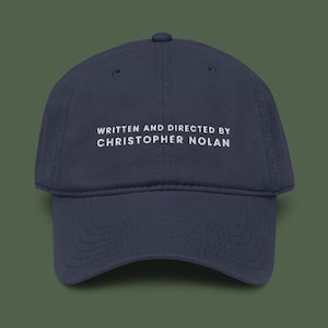 May include: A navy blue baseball cap featuring white embroidered text on the front that reads WRITTEN AND DIRECTED BY CHRISTOPHER NOLAN. The cap has a structured six-panel crown, a curved brim, and a button at the top centre of the crown.