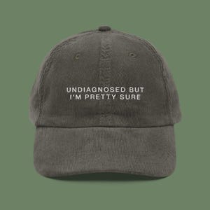May include: A dark green corduroy baseball cap with white embroidered text that reads "Undiagnosed but I'm pretty sure".