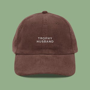 May include: A brown corduroy baseball cap with the text "TROPHY HUSBAND" embroidered in white on the front.
