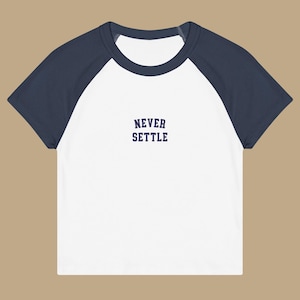 May include: White and navy blue baseball style t-shirt with the text "NEVER SETTLE" in navy blue block letters. The shirt has short sleeves and a crew neck.