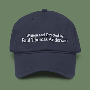 May include: A navy blue baseball cap featuring the embroidered text "Written and Directed by Paul Thomas Anderson" in white. The cap has a curved peak and a classic six-panel design, ideal for everyday use.