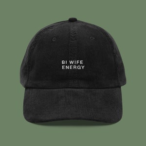 Peut inclure: Casquette de baseball en velours côtelé noir avec le texte brodé "BI WIFE ENERGY" en blanc. La casquette a une visière incurvée et un dessus arrondi. Le fond est vert olive uni.
