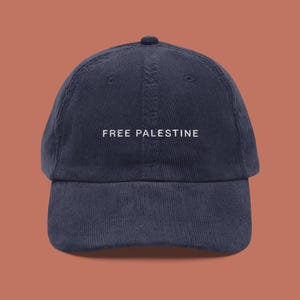 May include: Navy blue corduroy baseball cap with the text "FREE PALESTINE" embroidered in white across the front. The cap has a curved brim and a small button on top. The background is a solid shade of orange.