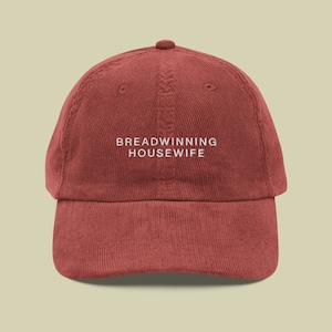 Op de afbeelding: Baksteenrode corduroy baseballcap met de witte geborduurde tekst "BREADWINNING HOUSEWIFE". De cap heeft een gebogen klep en een knoop aan de bovenkant.