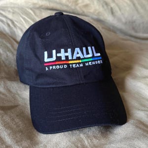 Puede incluir: Gorra de béisbol negra con el logotipo de U-HAUL bordado en blanco, rojo, naranja, amarillo, verde y azul. El texto "A PROUD TEAM MEMBER" está debajo. La gorra tiene una visera curva.
