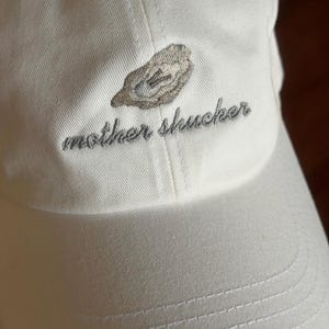 May include: A white baseball cap with the embroidered text "mother shucker" in gray script. Above the text is a gray embroidered oyster design. The cap is made of a soft fabric and has a curved brim.