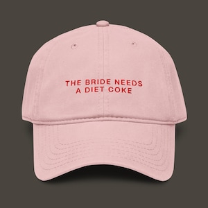 Op de afbeelding: Lichtroze baseballpet met een gebogen klep. De pet heeft de tekst "THE BRIDE NEEDS A DIET COKE" in rode letters op de voorkant. De pet is gemaakt van een zachte stof.