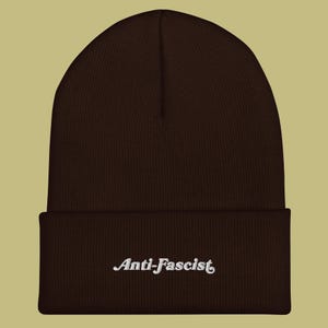 May include: A dark brown knit beanie featuring a cuffed brim. The front of the cuff displays the word Anti-Fascist embroidered in white thread using a stylised, cursive-inspired font. The hat has a ribbed texture throughout the crown and cuff.