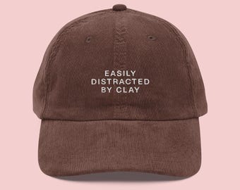 Może przedstawiać: Brązowa czapka bejsbolowa sztruksowa z białym napisem "EASILY DISTRACTED BY CLAY". Czapka ma zakrzywiony daszek i mały guzik na górze. Tło jest jasnoróżowe.