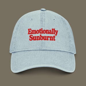 May include: Light blue denim baseball cap with the embroidered text "Emotionally Sunburnt" in red. The cap has a curved brim and a button on top. A casual accessory for everyday wear.