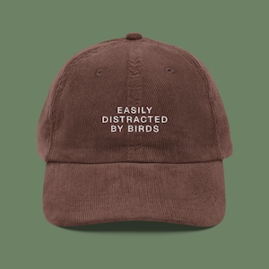 May include: A brown corduroy baseball cap with white embroidery that reads "EASILY DISTRACTED BY BIRDS".