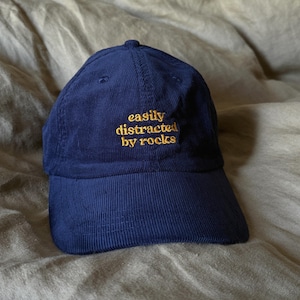 May include: Navy blue corduroy baseball cap with the embroidered text "easily distracted by rocks" in gold thread. The cap has a curved brim and a rounded top.