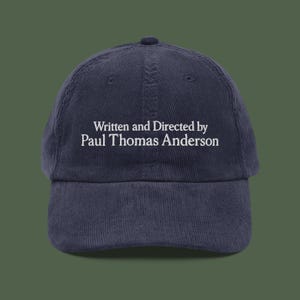 May include: A navy blue corduroy baseball cap featuring the white text "Written and Directed by Paul Thomas Anderson". The cap has a curved peak and a button on top. A classic style hat.