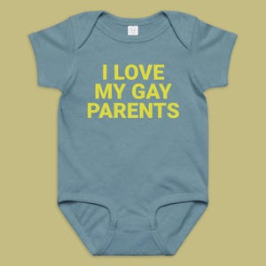 Può includere: Un body per neonati a maniche corte di colore blu polvere con il testo I LOVE MY GAY PARENTS stampato in grassetto giallo a lettere maiuscole sul petto. Il capo presenta uno scollo rotondo e aperture standard per le gambe dal design semplice.