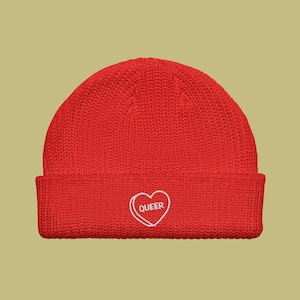 Puede incluir: Gorro de punto rojo con un corazón blanco bordado en la parte delantera. El corazón tiene la palabra "QUEER" en su interior.