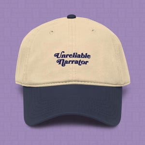 May include: A two-tone baseball cap featuring a beige crown and a dark navy blue brim. The front of the cap displays the text "Unreliable Narrator" embroidered in dark navy blue cursive lettering. The cap includes a small dark navy blue button on top.