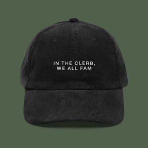 Puede incluir: Gorra de béisbol negra de pana con visera curva. La gorra presenta el texto blanco "IN THE CLERB, WE ALL FAM" centrado en la parte delantera. La gorra tiene un ajuste estructurado y un botón superior.