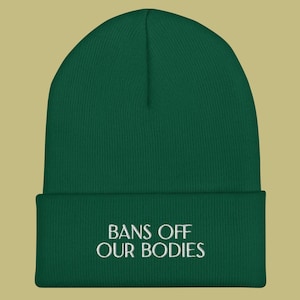 May include: A dark green cuffed beanie featuring the white embroidered text BANS OFF OUR BODIES centered on the front fold. The knit hat has a classic rounded crown and a thick ribbed texture, providing a simple and clean look for everyday wear.