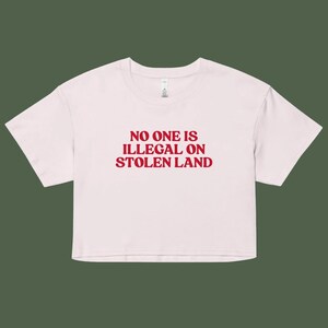 May include: A pale pink crop top featuring the red text "NO ONE IS ILLEGAL ON STOLEN LAND". The top has a round neckline and short sleeves. The material looks soft and comfortable.