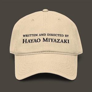 以下が含まれることがあります： ベージュ色の野球帽。黒字で「WRITTEN AND DIRECTED BY HAYAO MIYAZAKI」と書かれています。つばはカーブしており、上部にボタンが付いています。帽子は柔らかい生地で作られています。