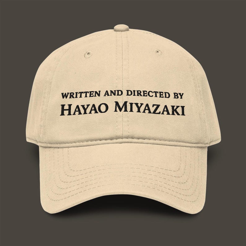 以下が含まれることがあります： ベージュ色の野球帽。黒字で「WRITTEN AND DIRECTED BY HAYAO MIYAZAKI」と書かれています。つばはカーブしており、上部にボタンが付いています。帽子は柔らかい生地で作られています。