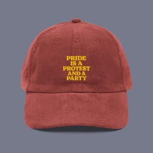 May include: A red corduroy baseball cap with the text "Pride is a protest and a party" embroidered in yellow.