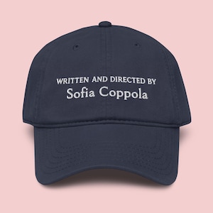 Può includere: Cappellino da baseball blu navy con testo ricamato bianco: "WRITTEN AND DIRECTED BY Sofia Coppola". Il cappellino ha una visiera curva e una chiusura con cinturino in tessuto.