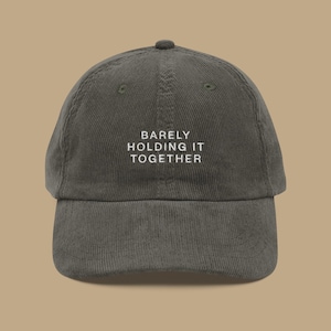 May include: A dark olive green corduroy baseball cap with a curved brim. The cap features the white embroidered text "BARELY HOLDING IT TOGETHER" centered on the front. The hat has a classic six-panel design with a top button.