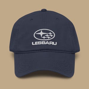 May include: Navy blue baseball cap with a white embroidered logo and the word "LESBARU". The logo features a stylized oval with stars. The cap has a curved brim and a structured crown.