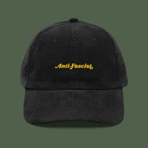 Könnte beinhalten: Schwarze Cord-Baseballkappe mit dem gelb gestickten Text "Anti-Fascist". Die Kappe hat einen gebogenen Schirm und eine strukturierte Krone. Der Hintergrund ist einfarbig olivgrün.