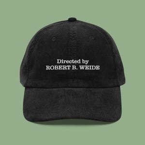 May include: Black corduroy baseball cap featuring the white embroidered text "Directed by ROBERT B. WEIDE". The cap has a curved peak and a button on top. A classic, casual accessory.