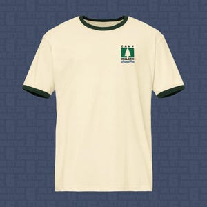 Pode incluir: Uma t-shirt ringer de cor creme com acabamentos verde-escuros na gola e nas mangas. O peito apresenta um pequeno gráfico com o texto CAMP WALDEN sobre um quadrado verde contendo um pinheiro branco e linhas onduladas azuis.