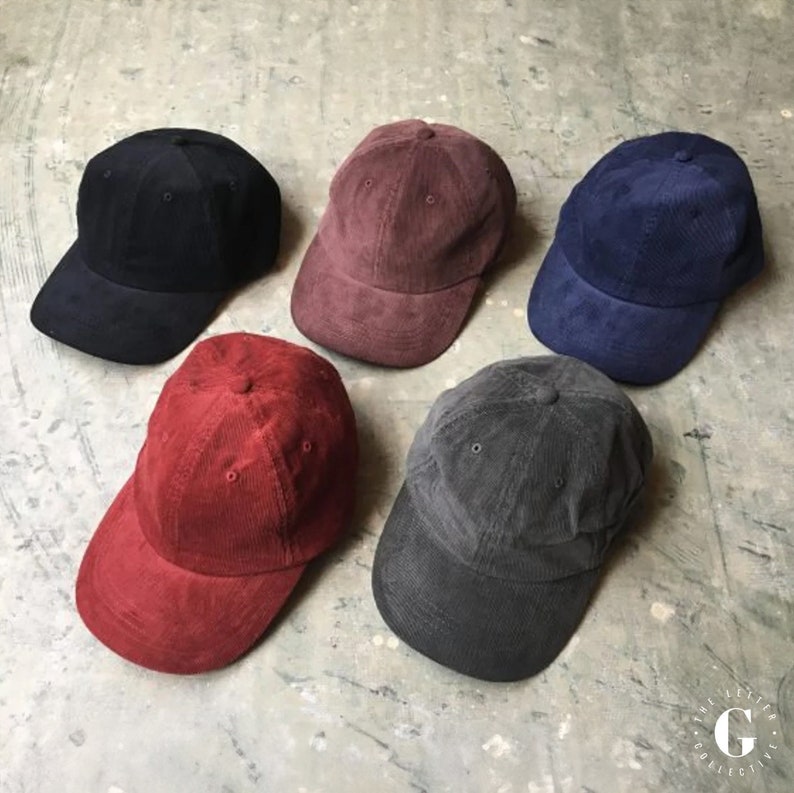 May include: Five corduroy baseball caps in different colours: black, burgundy, navy blue, red, and grey.